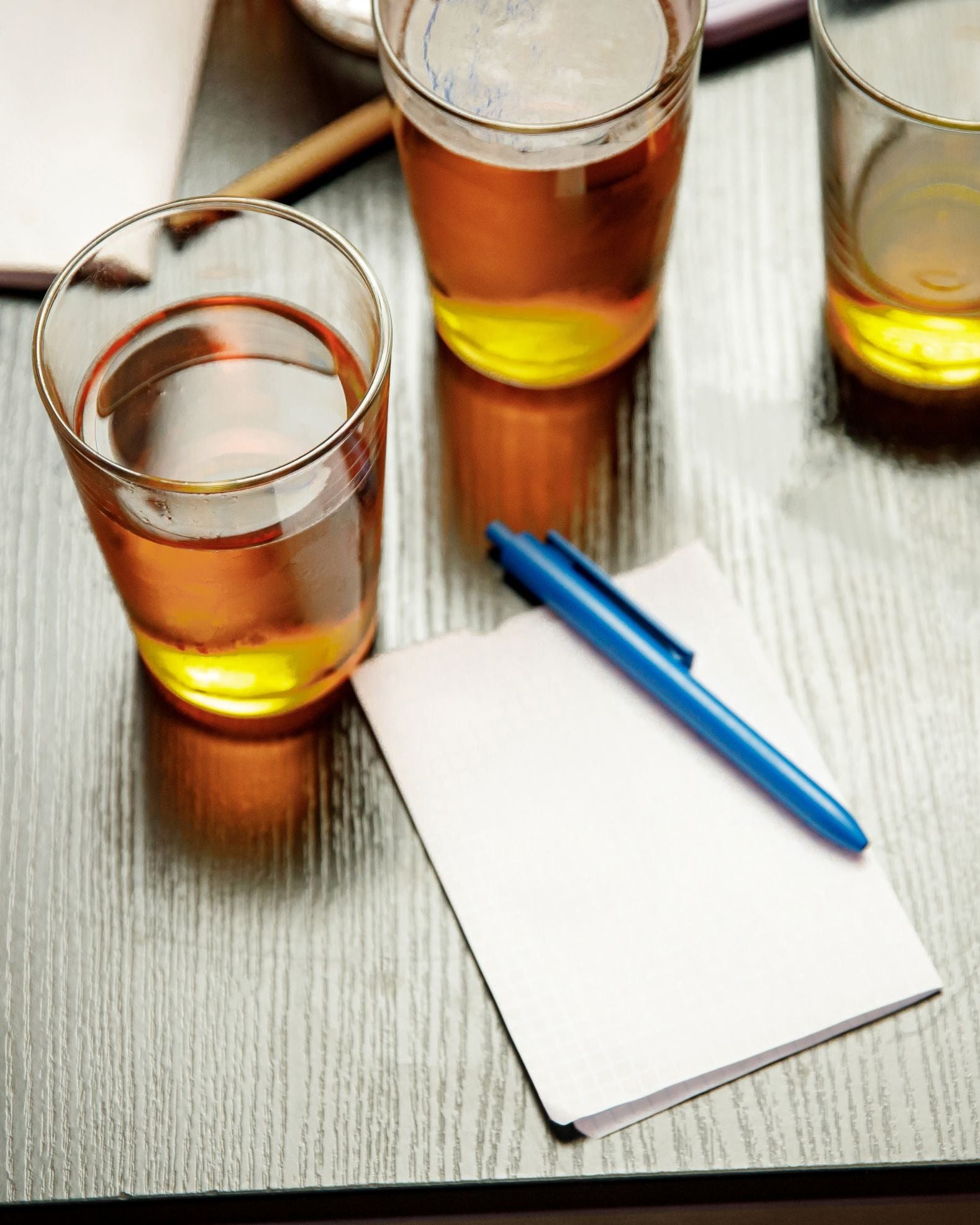 Three glasses of amber liquid on a wooden table with a pen and paper.